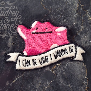 以下が含まれることがあります： 「I can be what I wanna be」というテキストが書かれた、ピンクと白の刺繍パッチ。漫画のキャラクターが描かれている。