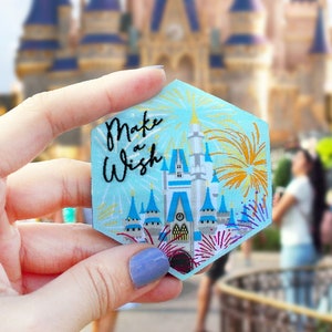 Puede incluir: Un parche hexagonal azul y dorado con una representación del castillo de Cenicienta y fuegos artificiales. El texto "Make a Wish" está escrito en letras cursivas blancas.