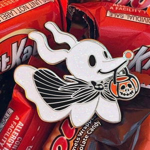 Puede incluir: Un pin de esmalte Zero blanco y brillante con un contorno dorado, de Pesadilla antes de Navidad. Zero sostiene un cubo de linterna de calabaza. El pin está sobre un fondo de barras de chocolate.