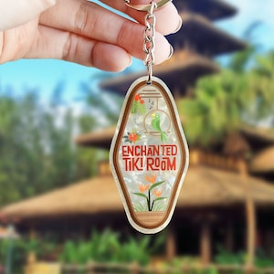 Puede incluir: Un llavero estilo motel con fondo blanco nacarado, que presenta un loro verde, flores tropicales naranjas y el texto rojo "ENCHANTED TIKI ROOM" impreso en el frente. El llavero está unido a una anilla y una cadena de metal plateado.