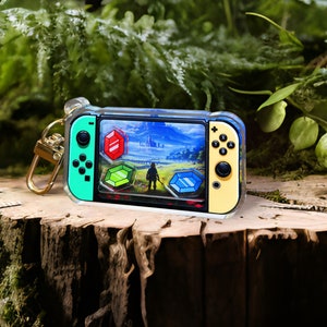 Puede incluir: Un llavero de acrílico transparente con forma de consola Nintendo Switch con un diseño verde y amarillo. La pantalla muestra una escena de videojuego con un personaje con una túnica azul y un paisaje de fondo. Hay tres gemas de colores en la pantalla.
