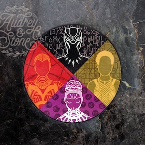 May include: A circular design with four sections, each featuring a different character from the Black Panther movie. The top section is black with a white silhouette of Black Panther. The right section is gold with a white silhouette of a woman wearing a necklace. The bottom section is purple with a white silhouette of a woman with a crown. The left section is red with a white silhouette of a woman wearing a necklace.