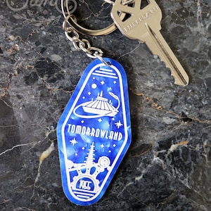 May include: A blue and white keychain with a Tomorrowland design. The keychain features a stylized image of the Space Mountain ride and the text "Tomorrowland" and "1955".