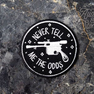 May include: A black and white embroidered patch with the text "NEVER TELL ME THE ODDS" encircling a white blaster pistol. The patch is round and has a white border. The background is a dark, marbled surface.