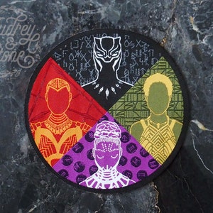 Pode incluir: Um remendo circular bordado com quatro seções. Cada seção apresenta um personagem diferente do filme Pantera Negra. A seção superior apresenta Pantera Negra em preto e branco. A seção direita apresenta Okoye em amarelo e verde. A seção inferior apresenta Shuri em roxo e branco. A seção esquerda apresenta Nakia em vermelho e laranja.