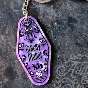 Puede incluir: Un llavero estilo motel de acrílico jaspeado en morado y blanco con un estampado gráfico negro de Gracey Manor, fantasmas y el número 999. El llavero está unido a una anilla de metal plateado con dos llaves de metal plateado.