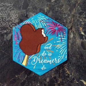 Pu&ograve; includere: Una toppa esagonale blu con un gelato Mickey Mouse marrone e fuochi d'artificio sullo sfondo. Il testo "and do as Dreamers do" &egrave; ricamato sulla toppa.