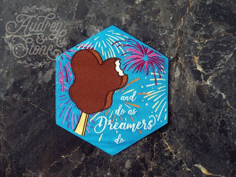 Pu&ograve; includere: Una toppa esagonale blu con un gelato Mickey Mouse marrone e fuochi d'artificio sullo sfondo. Il testo "and do as Dreamers do" &egrave; ricamato sulla toppa.