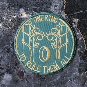 Pode incluir: Um remendo bordado verde com fio dourado que representa um anel com as palavras "Um Anel para governá-los a todos".