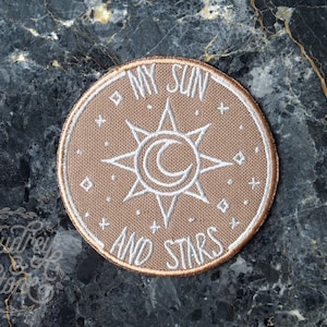 Può includere: Un cerotto in tessuto beige con ricamo bianco. Il cerotto presenta un sole con una luna crescente al suo interno e il testo "My Sun and Stars".