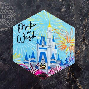 Pu&ograve; includere: Una toppa ricamata blu e bianca con una rappresentazione del Castello di Cenerentola con fuochi d'artificio sullo sfondo. Il testo "Make a Wish" &egrave; scritto in lettere corsive nere.