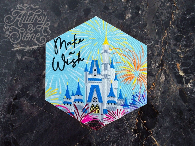 Pu&ograve; includere: Una toppa ricamata blu e bianca con una rappresentazione del Castello di Cenerentola con fuochi d'artificio sullo sfondo. Il testo "Make a Wish" &egrave; scritto in lettere corsive nere.