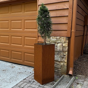 May include: A rusty brown metal planter with a small evergreen tree growing inside. The planter is sitting on a stone patio in front of a brown wooden garage door.