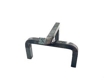 Puede incluir: Una herramienta de metal soldada de color gris oscuro con una forma única. Cuenta con una barra horizontal soportada por dos patas curvas y una barra vertical, todas conectadas en ángulo recto. La herramienta parece hecha a mano y tiene una estética industrial en bruto.