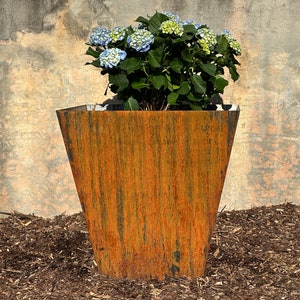May include: A rusty brown metal planter with a square top and a slightly flared base. A blue and white hydrangea plant is growing in the planter.