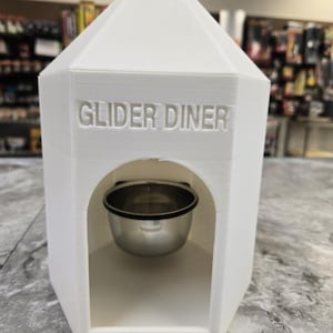 May include: A white, 3D-printed sugar glider diner with the words "GLIDER DINER" embossed on the front. The structure has a pentagonal roof and a rounded archway that houses a stainless steel bowl.
