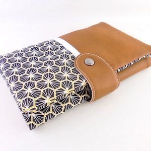 Peut inclure: Un portefeuille en cuir marron avec une section en tissu à motifs géométriques. Le tissu présente un motif noir, blanc et or. Le portefeuille a une fermeture à bouton-pression et des coutures blanches.