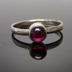 May include: A silver ring featuring a single, round, cabochon-cut deep magenta gemstone set in a smooth bezel setting. The band is polished silver with a simple, rounded profile, showcasing a reflective surface and a clear, vibrant stone.