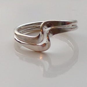 Può includere: Un anello d'argento con un design unico a forma di onda. L'anello presenta una finitura liscia e lucida e una struttura a doppia fascia, creando un look moderno ed elegante. Le curve dell'anello riflettono la luce.