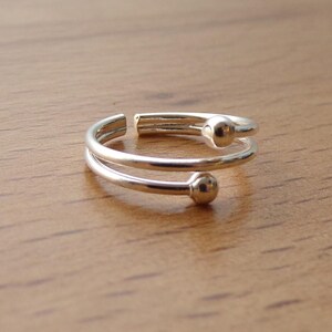 May include: A silver-toned bypass ring featuring a double-band design with an open gap. Each end of the band terminates in a small, smooth, spherical ball accent. The metal has a polished, reflective finish that catches the light.