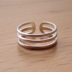 Pode incluir: Um anel de dedo do pé prateado com três faixas paralelas. O design aberto permite um ajuste regulável. O anel é feito de metal brilhante e assenta numa superfície de madeira castanha clara.
