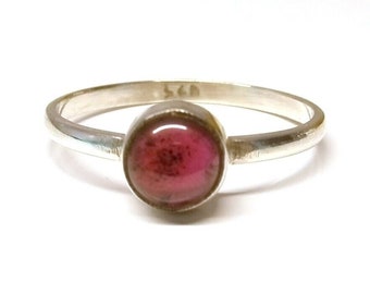 Puede incluir: Anillo de plata con una piedra preciosa rosa redonda. El anillo tiene una banda simple y un engaste de bisel. La piedra preciosa tiene un aspecto moteado, con inclusiones rosas más oscuras. Joya delicada.