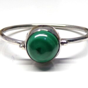 May include: A silver ring featuring a smooth, round, polished malachite gemstone set in a simple metal bezel. The band is thin and circular, showcasing a vibrant green stone with natural dark green banding patterns centered on the setting.