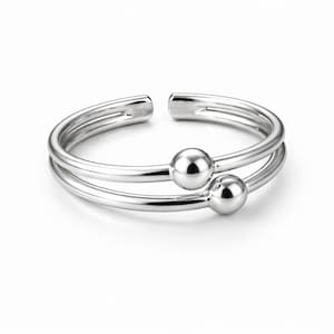 Könnte beinhalten: Ein silberfarbener Zehenring mit Doppelband-Design. Der Ring hat zwei kleine, runde, polierte Kugeln und ein offenes Design. Der Ring besteht aus glänzendem, reflektierendem Metall und ist vor weißem Hintergrund.