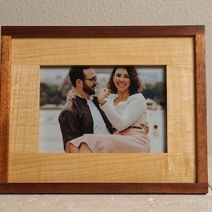 May include: A wooden picture frame with a light brown wood border and a darker brown wood trim. The frame holds a photo of a couple smiling and looking at each other.