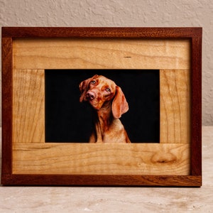 May include: A wooden picture frame with a reddish-brown outer frame and a lighter wood inner frame. A photo of a brown dog with its head tilted to the side is inside the frame.