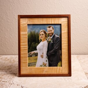 May include: A wooden picture frame with a natural wood finish and a red mahogany border. The frame holds a photo of a bride and groom standing together in a field.