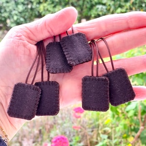 Könnte beinhalten: Eine Ansammlung von fünf kleinen, rechteckigen dunkelbraunen Filzanhängern, die jeweils einen genähten Rand und eine dünne, oben befestigte braune Kordelschlaufe aufweisen. Die Anhänger sind in Form und Farbe einheitlich und haben eine weiche Oberfläche.