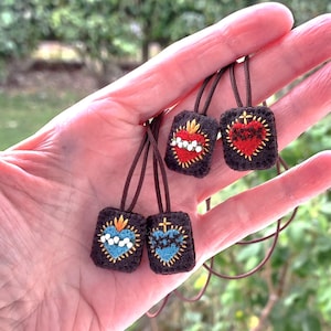 Peut inclure: Quatre petits pendentifs rectangulaires avec des motifs de cœur brodés en rouge, bleu et or. Chaque pendentif est attaché à un cordon marron. Les pendentifs sont tenus dans une main sur un fond vert flou.