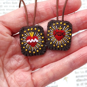 May include: Two rectangular dark brown felt pendants featuring embroidered sacred heart designs in red, white, and yellow thread. Each pendant has a brown cord attached at the top, with radiating sunburst patterns and small yellow dots surrounding the heart.