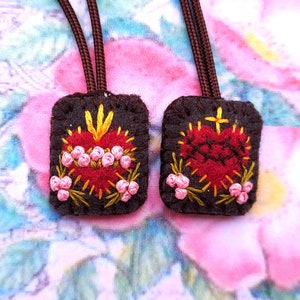 May include: Two rectangular dark brown felt patches featuring embroidered red hearts. One heart is adorned with pink roses and yellow flames, while the other features a crown of thorns and a yellow cross. Each patch has a brown cord attached at the top.