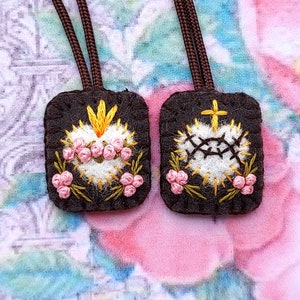 May include: Two rectangular dark brown felt scapulars featuring hand-embroidered white hearts with pink rose accents. The left heart has yellow flames, while the right heart features a black crown of thorns and a yellow cross. Each has a brown cord.