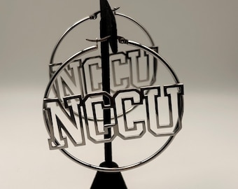 May include: A pair of silver-toned hoop earrings featuring the letters NCCU in a block font integrated into the center of each circular frame. The earrings are constructed from a polished metallic material with a clear, open-work design.