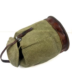 Peut inclure: Un sac en toile vert olive avec une garniture et des sangles en cuir marron. Le sac a une fermeture à cordon et une sangle en cuir avec une boucle en métal. Le sac est incliné, montrant le côté et le dessus.