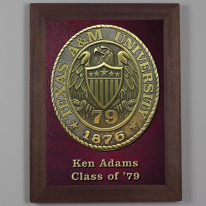 May include: A framed plaque featuring a metallic seal of Texas A&M University. The seal includes an eagle, stars, and the year 1876. The plaque is framed in dark wood and has a burgundy background with the name "Ken Adams" and "Class of '79".