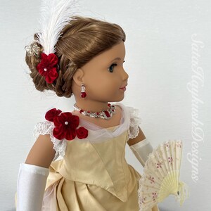 Puede incluir: Una mu&ntilde;eca con un vestido color crema con detalles florales de terciopelo rojo, un collar a juego, aretes y un abanico. El cabello de la mu&ntilde;eca est&aacute; peinado con una pluma blanca y accesorios para el cabello florales rojos. La mu&ntilde;eca lleva guantes blancos.
