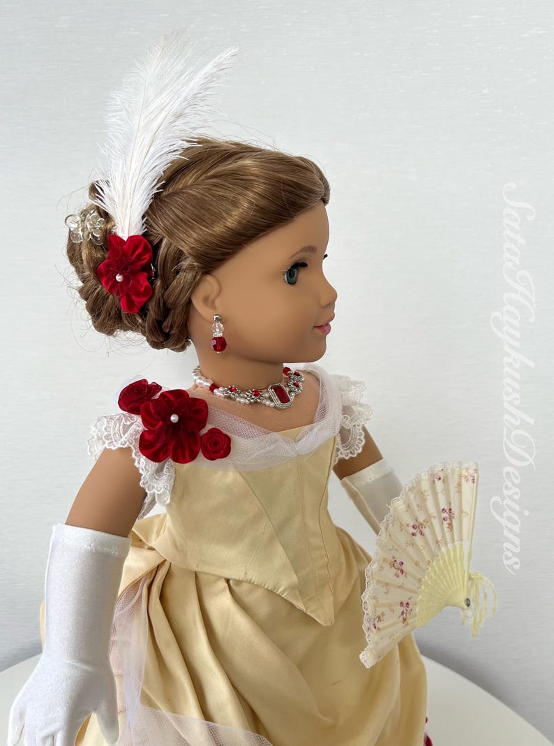 Puede incluir: Una mu&ntilde;eca con un vestido color crema con detalles florales de terciopelo rojo, un collar a juego, aretes y un abanico. El cabello de la mu&ntilde;eca est&aacute; peinado con una pluma blanca y accesorios para el cabello florales rojos. La mu&ntilde;eca lleva guantes blancos.