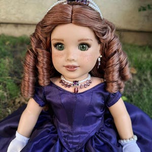 Puede incluir: Mu&ntilde;eca con cabello casta&ntilde;o rojizo y ojos verdes, vestida con un vestido de sat&eacute;n morado oscuro. La mu&ntilde;eca est&aacute; adornada con un collar, pendientes y una tiara de perlas y amatistas. Guantes blancos y una pulsera de perlas completan el conjunto.