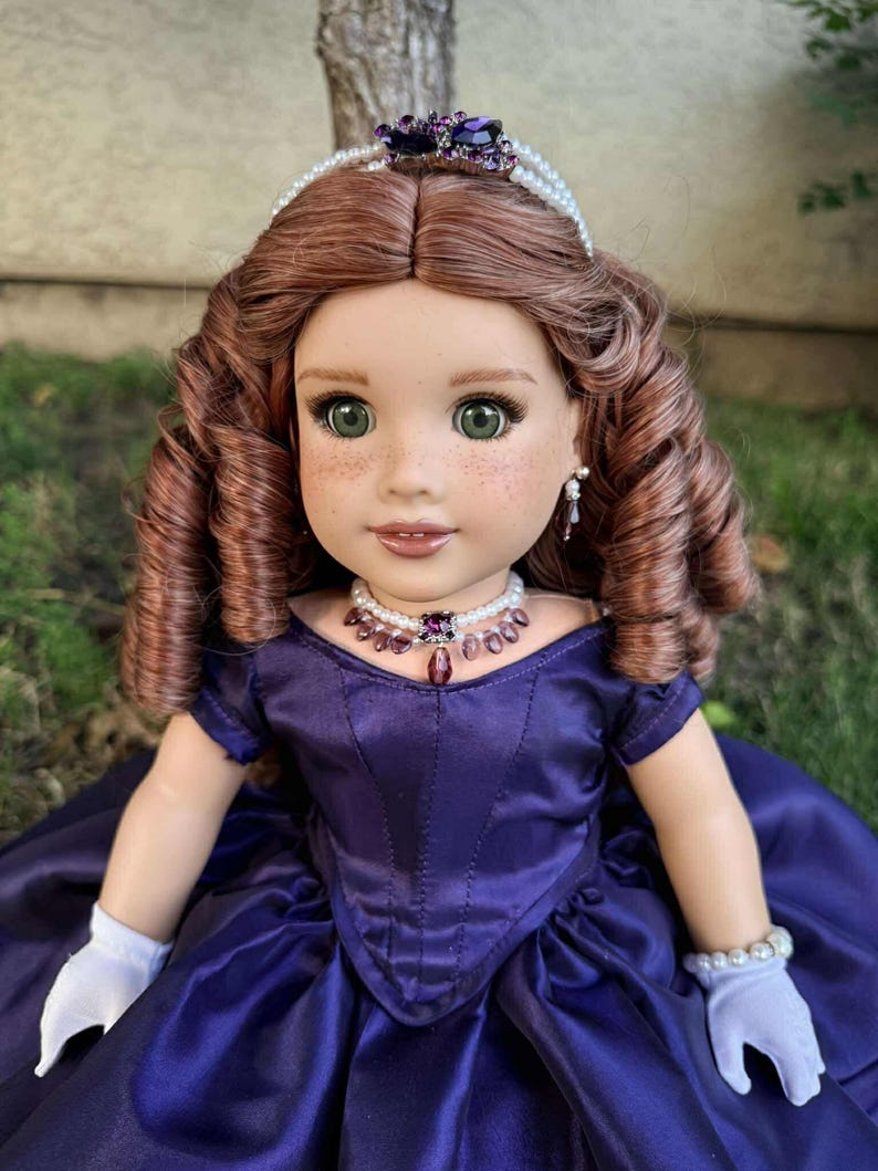 Puede incluir: Mu&ntilde;eca con cabello casta&ntilde;o rojizo y ojos verdes, vestida con un vestido de sat&eacute;n morado oscuro. La mu&ntilde;eca est&aacute; adornada con un collar, pendientes y una tiara de perlas y amatistas. Guantes blancos y una pulsera de perlas completan el conjunto.