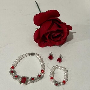 Puede incluir: Un conjunto de joyas con pulsera, anillo y pendientes con perlas de imitaci&oacute;n y detalles rojos. La pulsera tiene una gran piedra roja rectangular en el centro, y los pendientes y el anillo tienen cuentas rojas a juego. Tambi&eacute;n hay una rosa roja.