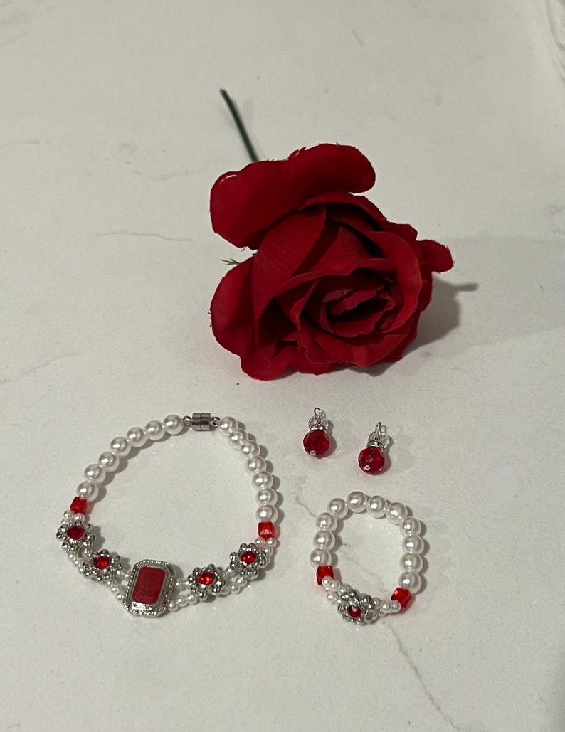 Puede incluir: Un conjunto de joyas con pulsera, anillo y pendientes con perlas de imitaci&oacute;n y detalles rojos. La pulsera tiene una gran piedra roja rectangular en el centro, y los pendientes y el anillo tienen cuentas rojas a juego. Tambi&eacute;n hay una rosa roja.