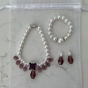 Puede incluir: Un conjunto de collar, pulsera y pendientes de perlas con detalles morados. El collar presenta una gema cuadrada morada y cuentas moradas en forma de l&aacute;grima. La pulsera y los pendientes tambi&eacute;n tienen gemas moradas.