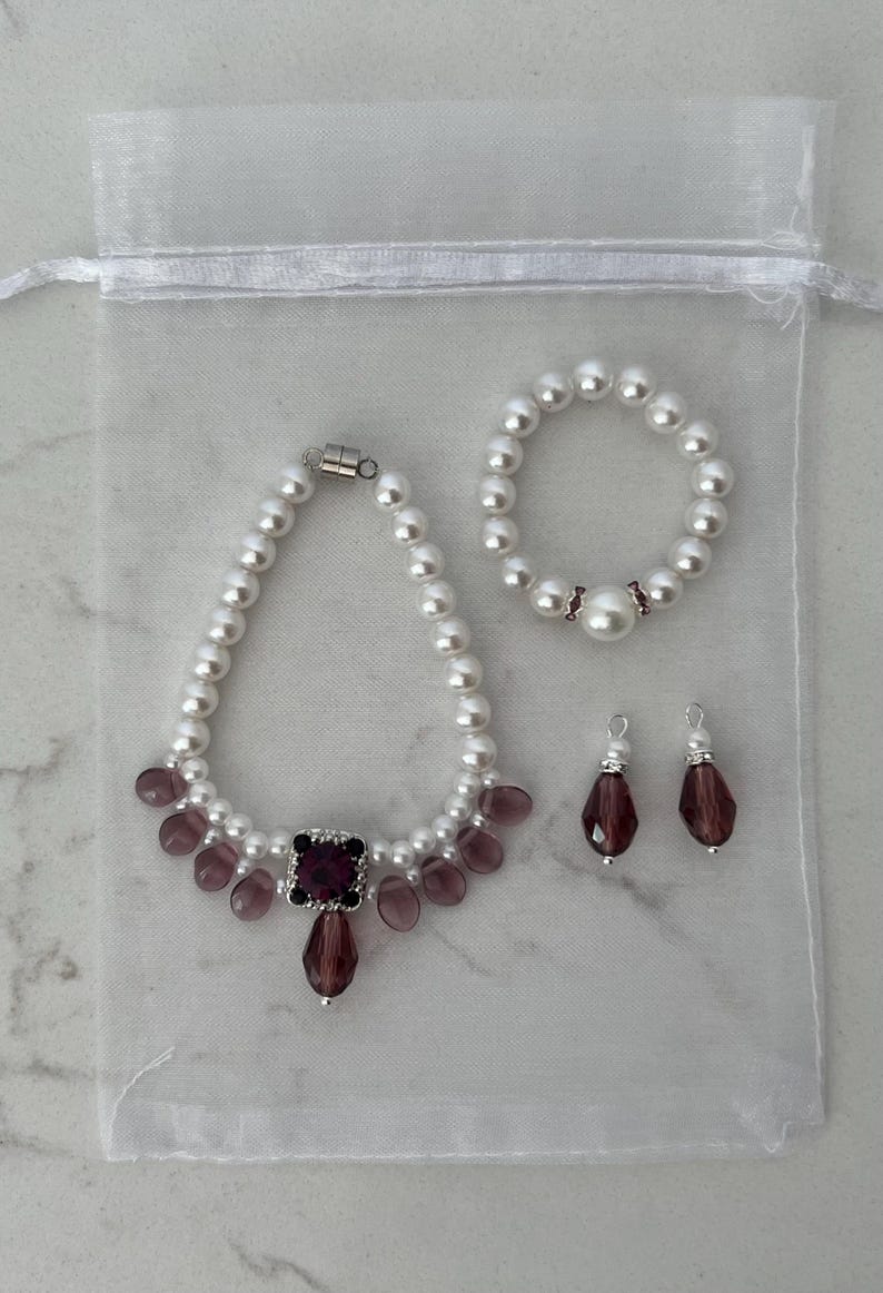 Puede incluir: Un conjunto de collar, pulsera y pendientes de perlas con detalles morados. El collar presenta una gema cuadrada morada y cuentas moradas en forma de l&aacute;grima. La pulsera y los pendientes tambi&eacute;n tienen gemas moradas.