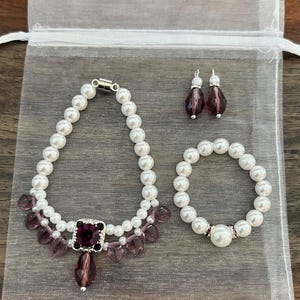 Puede incluir: Un conjunto de joyas con collar, pulsera y pendientes. El collar y la pulsera est&aacute;n hechos de perlas blancas y est&aacute;n acentuados con cuentas moradas y una gran gema morada cuadrada. Los pendientes tienen la parte superior plateada y cuentas moradas en forma de l&aacute;grima.