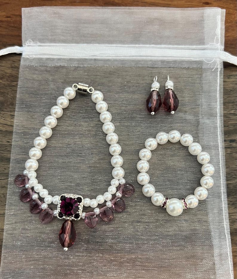 Puede incluir: Un conjunto de joyas con collar, pulsera y pendientes. El collar y la pulsera est&aacute;n hechos de perlas blancas y est&aacute;n acentuados con cuentas moradas y una gran gema morada cuadrada. Los pendientes tienen la parte superior plateada y cuentas moradas en forma de l&aacute;grima.