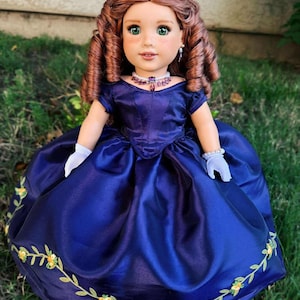 Puede incluir: Mu&ntilde;eca con cabello casta&ntilde;o rojizo y ojos verdes, vestida con un vestido de sat&eacute;n azul oscuro con bordados florales. La mu&ntilde;eca lleva una tiara y un collar, y est&aacute; sobre hierba verde.